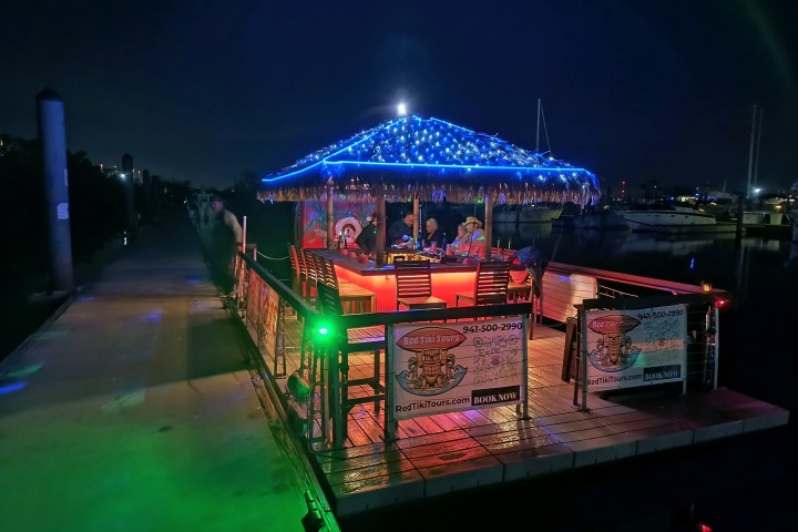 Floating tiki hut bar with glowing lights docked at night with people seated inside.