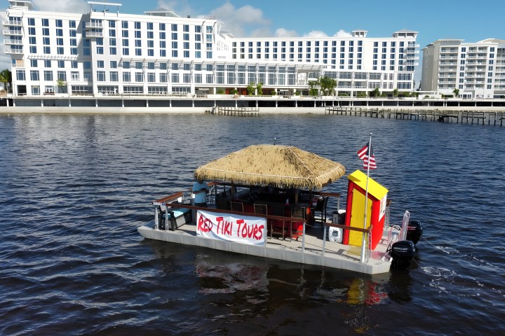 Small tiki bar boat with straw roof and American flag on water, large building in background.