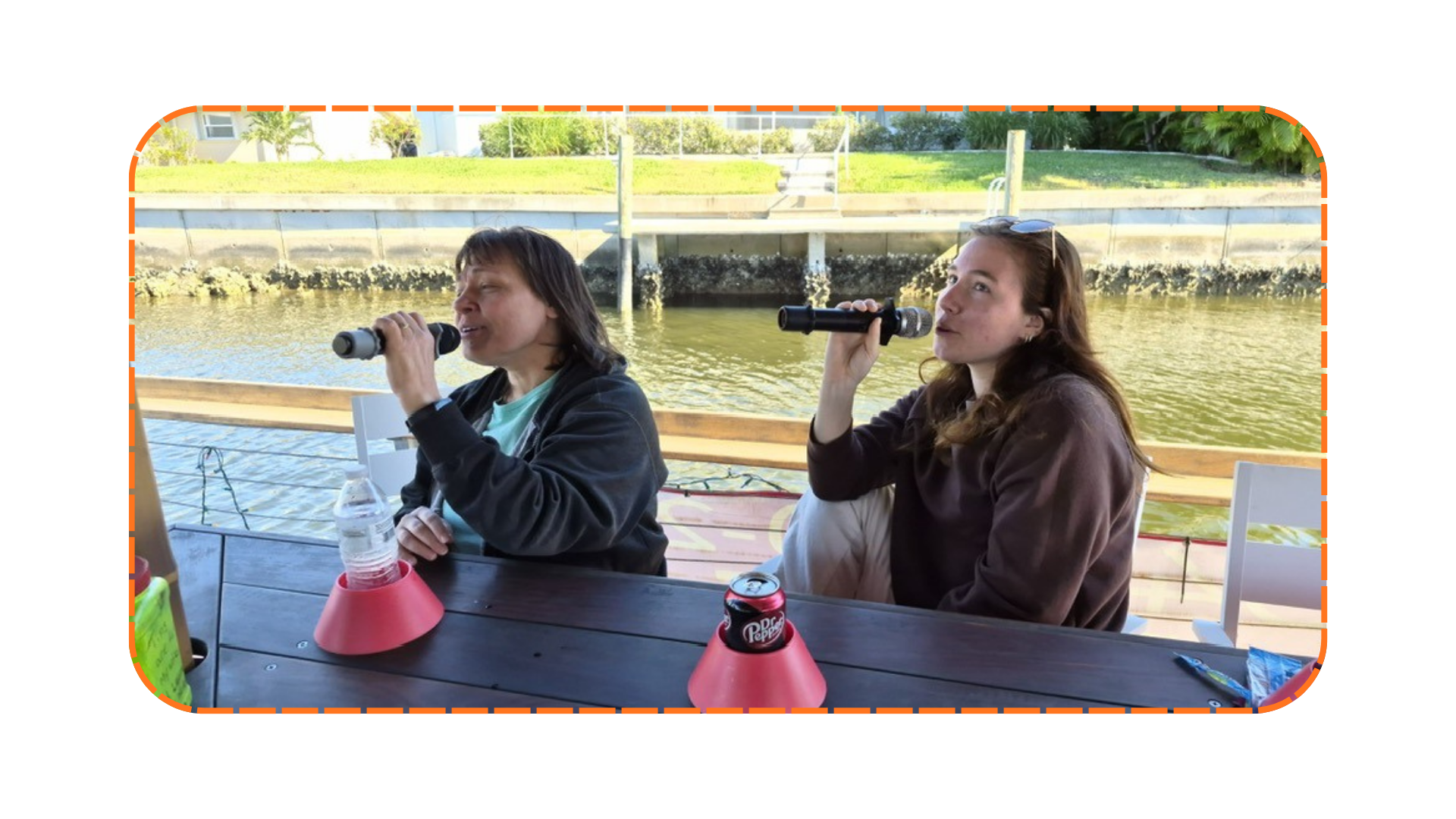 Two people seated by water, singing into microphones at a wooden table with drinks.