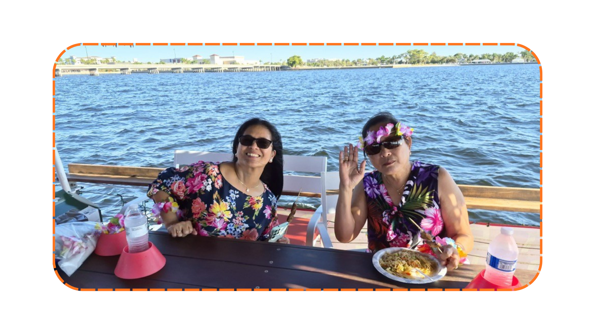 Two people in floral clothes sitting by a lake, with food on the table.