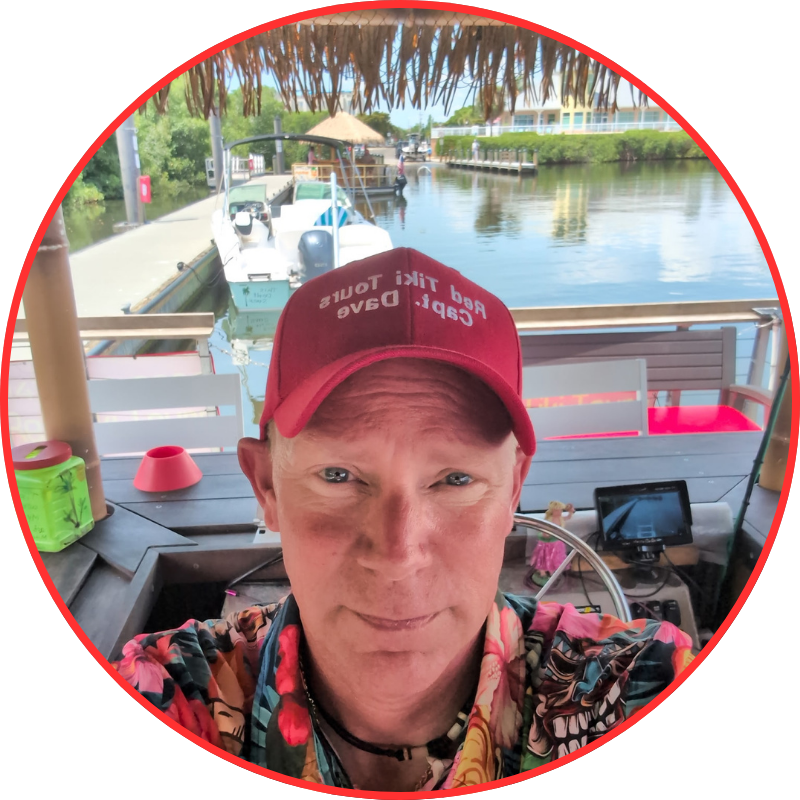 Person in floral shirt and red cap on a boat with a scenic waterfront backdrop.